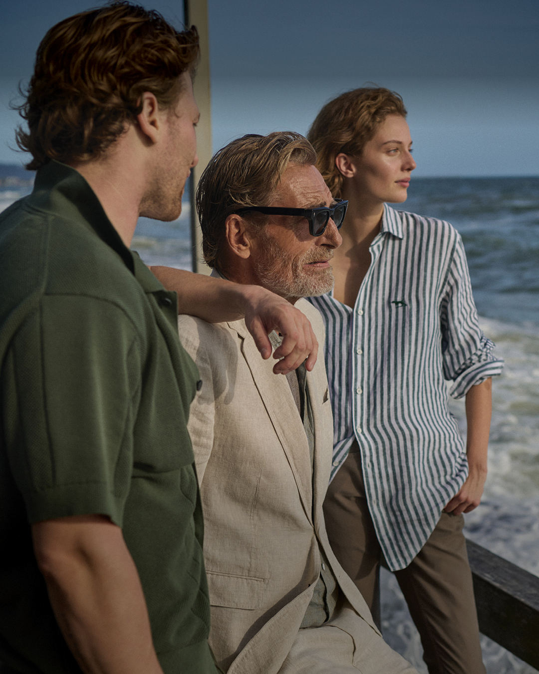 Three people standing on a seaside deck beside choppy waves, wearing an olive short-sleeve shirt, a beige blazer, and a blue-and-white striped button-down